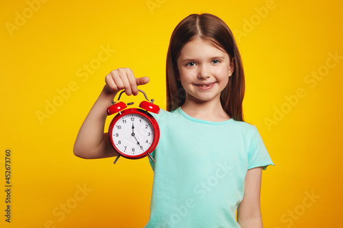 Adorable little kid girl in casual dress smiling and holding alarm clock against yellow studio background photo