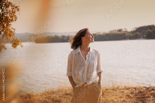 Beautiful young relaxed woman in white blouse enjoying nature breathing fresh air meditating on the river on an autumn day photo