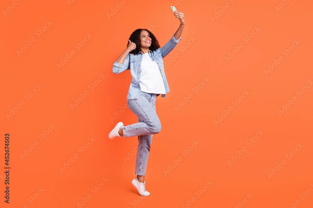 Full size photo of funky young brunette lady do selfie thumb up wear jeans shirt shoes isolated on orange background