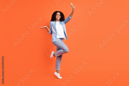 Full size photo of cool young brunette lady do selfie wear jeans shirt shoes isolated on orange color background