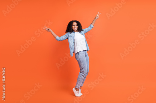 Full body photo of cool young brunette lady show v-sign wear jeans shirt shoes isolated on orange background
