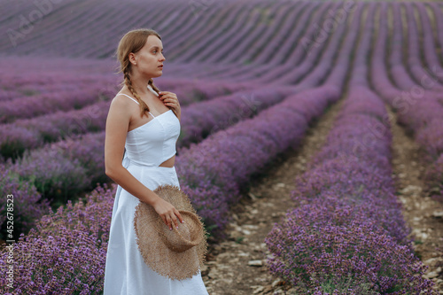A beautiful young woman in a white dress with a hat walks through a lavender field