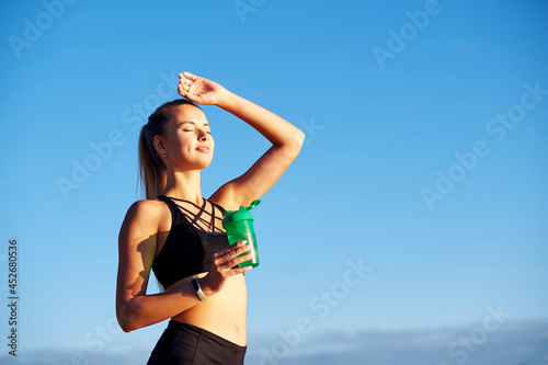 Fitness woman with bottle of water after running training on blue sky background
