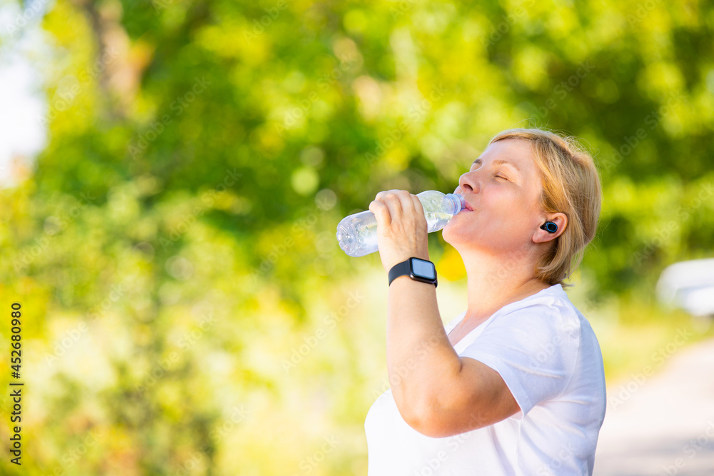 Beautiful sporty blonde woman wearing headphones and digital smart watch has a break after jogging outdoors in the park, drinking water from the plastic bottle. Beautiful sporty blonde woman wearing headphones and digital smart watch has a break after jogging outdoors in the park, drinking water from the plastic bottle.