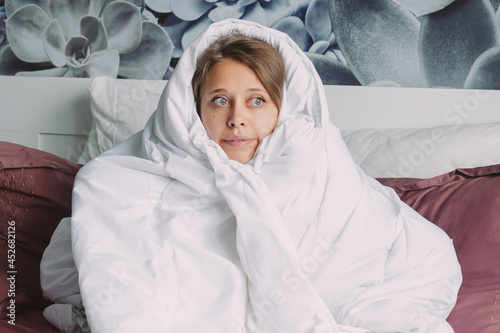 Vászonkép A young frozen funny caucasian charming blonde woman hides under a large white warm blanket to escape from the cold sitting on the bed in her room