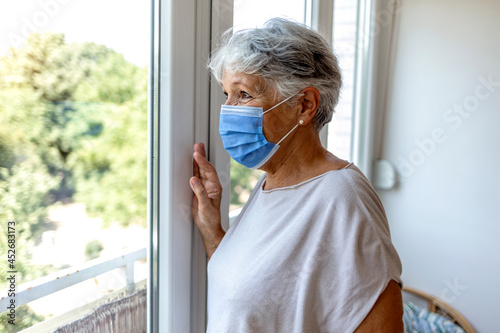 Portrait of beautiful senior woman wearing face mask and standing near window at home during the day. Old woman with grey hair wearing surgical mask and looking through the window during the lockdown.