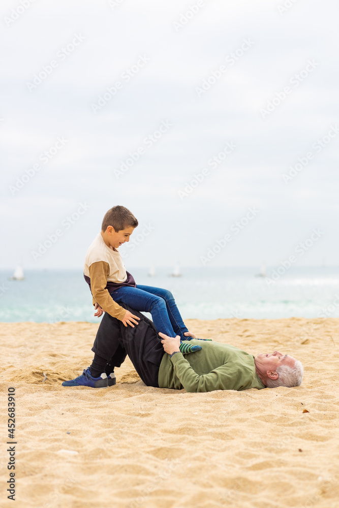 Funny boy and his grandfather playing stretched out in the sand on the beach