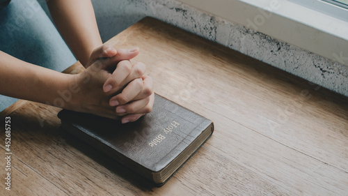 Woman with Bible praying, hands clasped together on her Bible on wooden table.