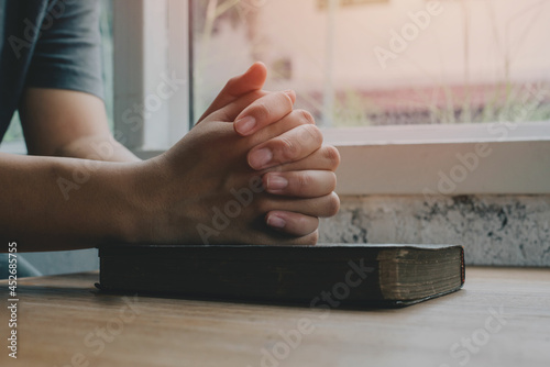 Woman with Bible praying, hands clasped together on her Bible on wooden table.