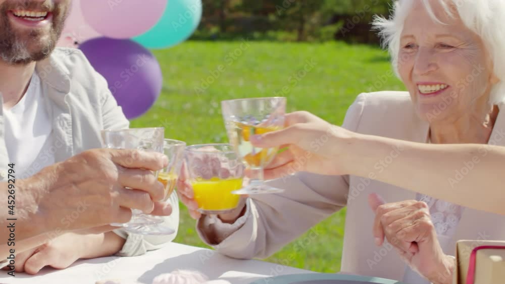 Close up shot of senior grandmother in party hat toasting with glass of orange juice, smiling and drinking during outdoor birthday celebration with family on sunny summer day Close up shot of senior grandmother in party hat toasting with glass of orange juice, smiling and drinking during outdoor birthday celebration with family on sunny summer day