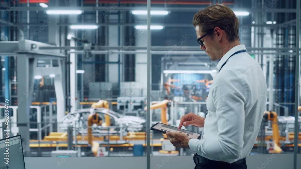 Handsome Engineer in Glasses and White Shirt Using Tablet Computer and Looking Out of the Office at a Car Assembly Plant. Industrial Specialist Working on Vehicle Design in Technological Facility. Handsome Engineer in Glasses and White Shirt Using Tablet Computer and Looking Out of the Office at a Car Assembly Plant. Industrial Specialist Working on Vehicle Design in Technological Facility.