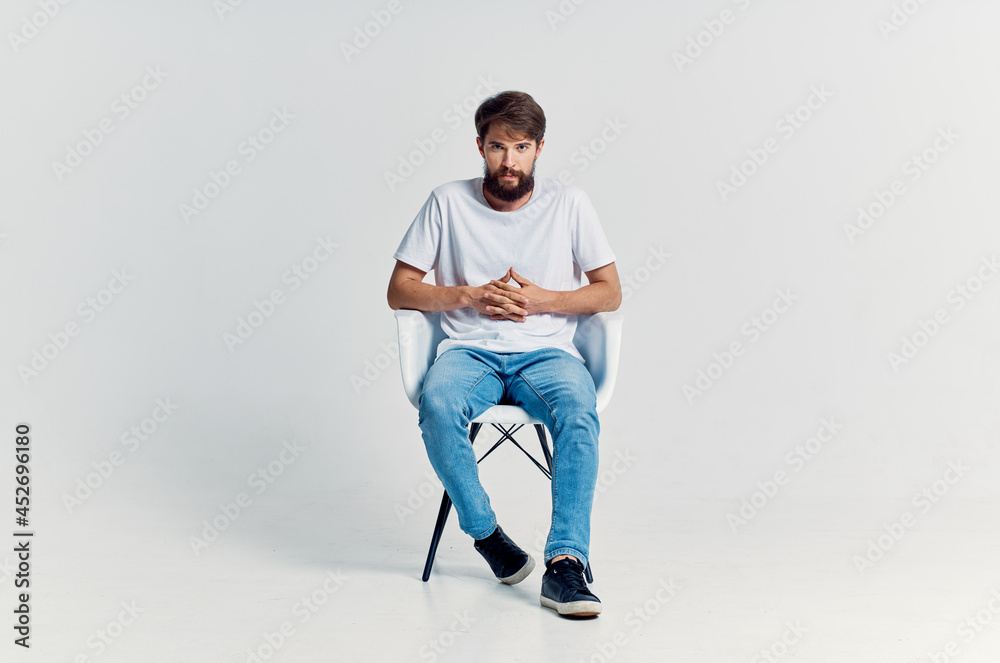 bearded man in white t-shirt sits on a chair light background