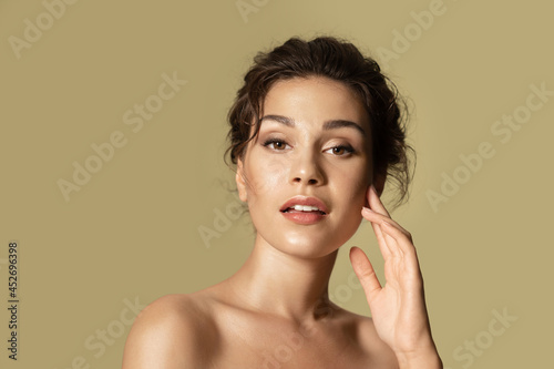 Pretty young female model with fresh healthy glowing skin, modern make up, bare shoulders. Confident girl posing on beige olive studio background with copy space. Natural woman beauty and femininity.
