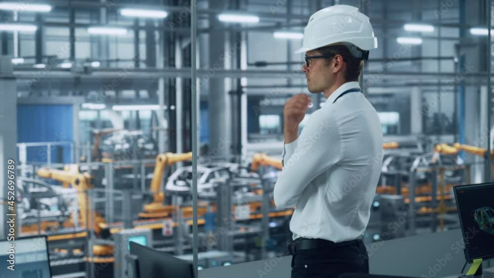 Handsome Engineer in Glasses and White Hard Hat Looking Out of the Office Window at a Car Assembly Plant. Industrial Specialist Working on Vehicle Production in Technological Manufacturing Facility. Handsome Engineer in Glasses and White Hard Hat Looking Out of the Office Window at a Car Assembly Plant. Industrial Specialist Working on Vehicle Production in Technological Manufacturing Facility.