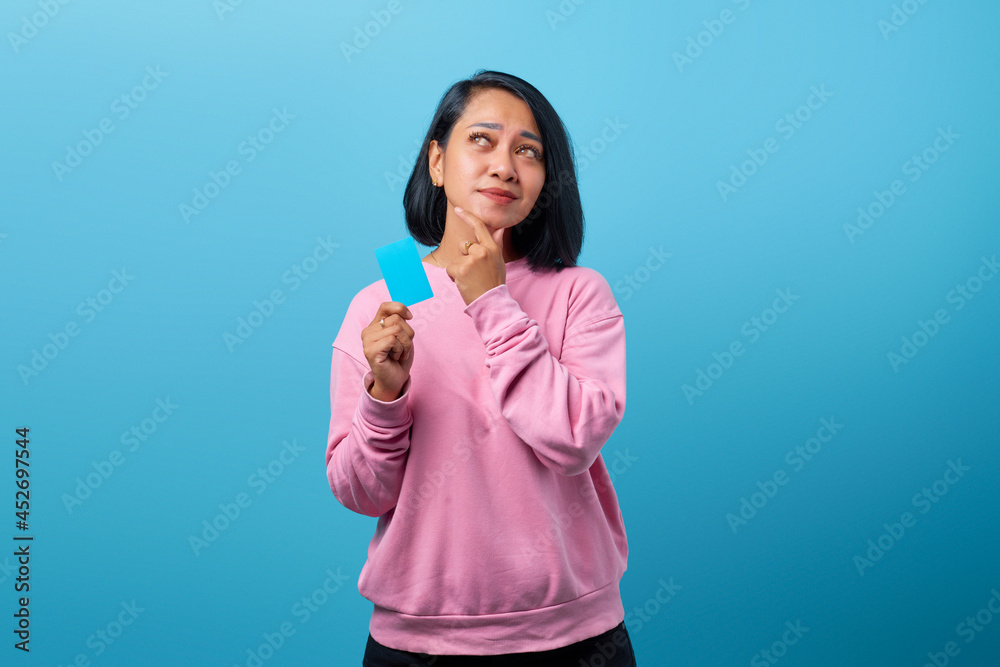 Portrait of young Asian woman holding credit card looking sideways with doubtful and skeptical expression Portrait of young Asian woman holding credit card looking sideways with doubtful and skeptical expression