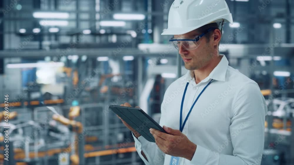 Male Engineer in Safety Goggles and Hard Hat Using Tablet Computer and Looking Out of the Office at a Car Assembly Plant. Industrial Specialist Working on Vehicle Production in Technological Factory. Male Engineer in Safety Goggles and Hard Hat Using Tablet Computer and Looking Out of the Office at a Car Assembly Plant. Industrial Specialist Working on Vehicle Production in Technological Factory.