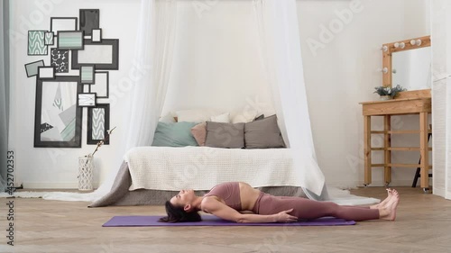 Slender, flexible, caucasian middle-aged woman with black hair in beige tracksuit performs standing asanas Raised Legs Pose uttana padasana on purple yoga mat in bright room with bed, mirror, window photo