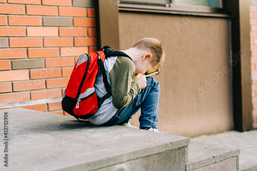 Back to school. Cute child with backpack, holding notepad and training books. School boy pupil with bag. Elementary school student going to classes. Tired Kid sitting on stairs outdoors at brick wall.