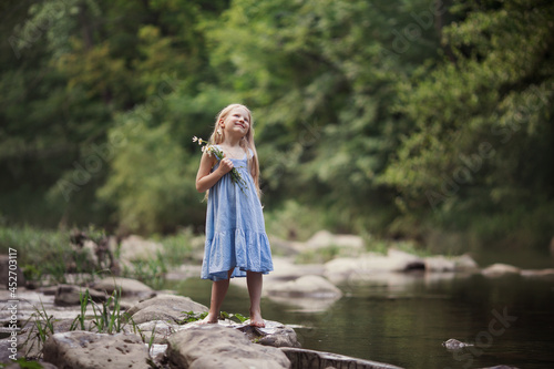 Fototapeta Naklejka Na Ścianę i Meble -  a little girl in a blue sundress with a bouquet of daisies walks by the river splashes of water