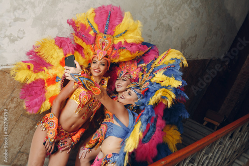 Woman in brazilian samba carnival costume with colorful feathers plumage with mobile phone take selfie in old entrance with big window.