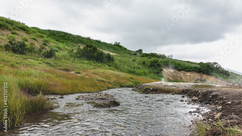 A stream flows in the valley of hot springs. There are sulfur deposits on the soil of the shores. Steam and haze above the ground. Green vegetation on the hillside. Kamchatka