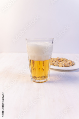 Glass of light beer with foam and plate with pistaches on white background