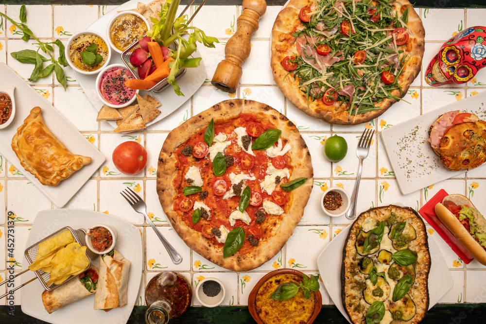 Top view image of international food dishes. Pizza with buffalo cheese and basil, hummus of various colors, burritos, Argentine empanadas, roasted aubergine, tomatoes and limes