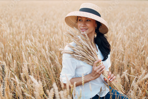 Young Woman in straw hat holding sheaf of wheat ears at agricultural field.