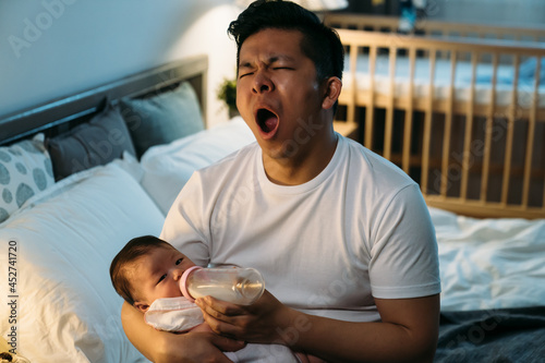 tired asian first time dad is yawning while sitting at bedside feeding his newborn girl bottled milk late at night in the bedroom at home.