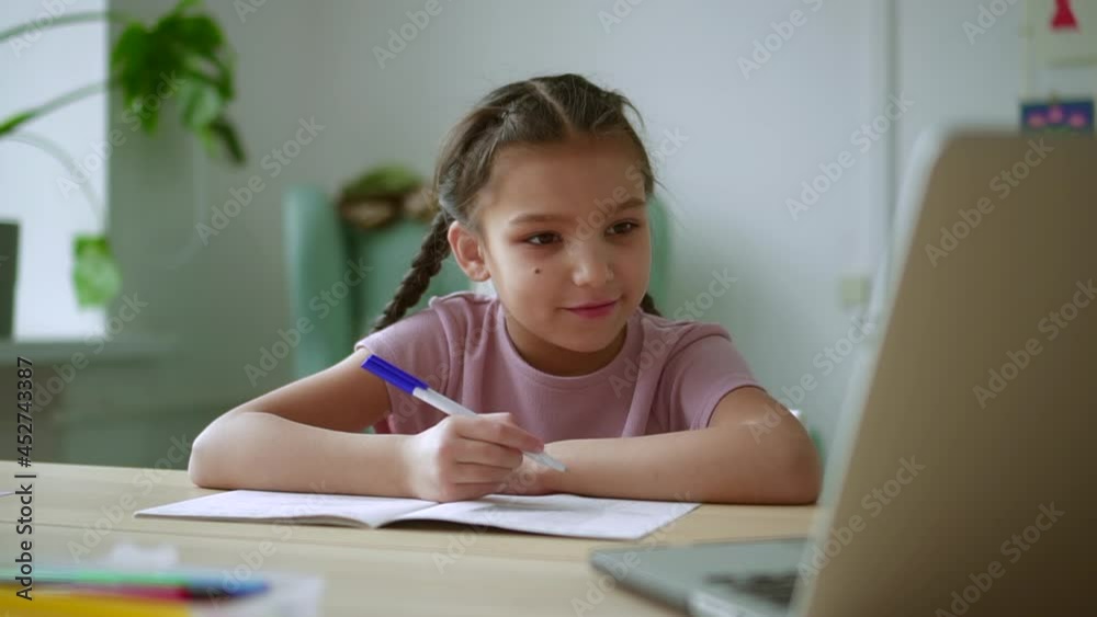 Home education for children. Spbd Attentive brunette girl pupil writes in exercise book listening to teacher explanation at video lesson in light room Home education for children. Spbd Attentive brunette girl pupil writes in exercise book listening to teacher explanation at video lesson in light room