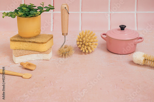 Natural eco friendly bamboo cleaning brushes, sponges, kitchen utensils, casserole dish and a green plant in a yellow pot on a pink tile background.