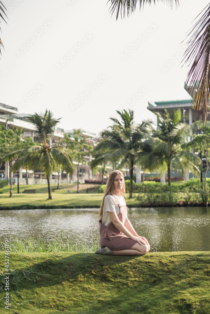 Pretty girl in casual clothing sitting the grass in the park. Beautiful sunshine. Long blond hair. Caucasian young woman. Teenage girl