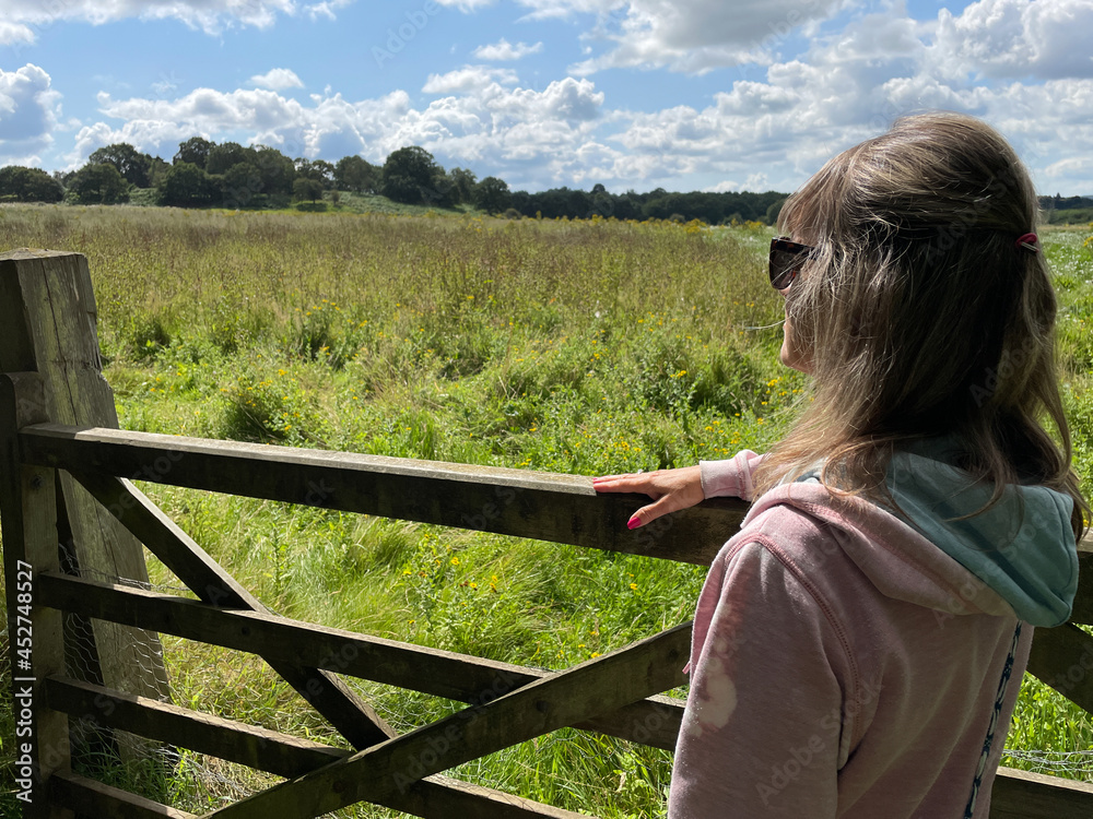 A lady in a pink hoody wearing sunglasses leans on a wooden farm gate looking out towards a wild meadow in background.Sunny day with blue sky and white clouds