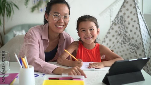 Happy family studying online and posing for camera while sitting at table in home room spbd. Front view of mom, daughter pose and look with smile, sit at desk in interior, girl writes in notebook photo