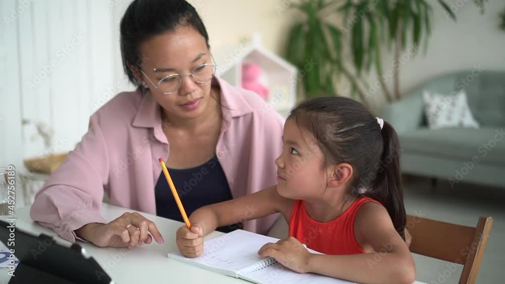 Online distance: woman helping girl to study and sitting at table in front of tablet in room spbd. Young mother teaches daughter and talks, cute girl writes in notebook and looks at device screen Online distance: woman helping girl to study and sitting at table in front of tablet in room spbd. Young mother teaches daughter and talks, cute girl writes in notebook and looks at device screen