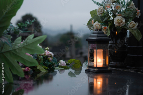 The lantern with candle and flowers as a decoration on the marble grave and cemetery in the background.