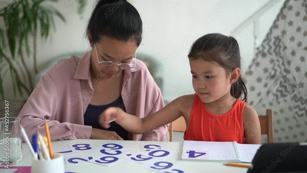 Online primary school education. Spbd Smart little Asian girl student makes number of cards with digits solving sums with tutor near tablet in light room Online primary school education. Spbd Smart little Asian girl student makes number of cards with digits solving sums with tutor near tablet in light room
