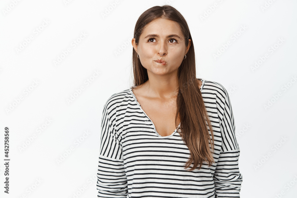 Image of woman thinking, pucker lips and looking up as if making choice, deciding, standing pensive against white background Image of woman thinking, pucker lips and looking up as if making choice, deciding, standing pensive against white background