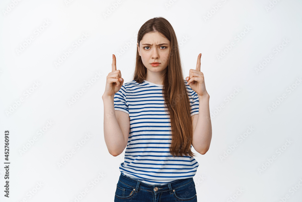 Upset gloomy girl frowning, pointing fingers up at empty space, standing in casual clothes against white background Upset gloomy girl frowning, pointing fingers up at empty space, standing in casual clothes against white background
