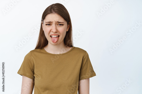 Young woman stick tongue from dislike, disgusted with bad taste, smth awful or disgusting, standing over white background photo