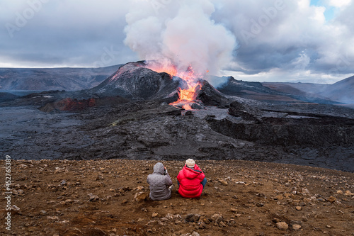 Faceless tourists contemplating active volcano from mountain under cloudy sky photo