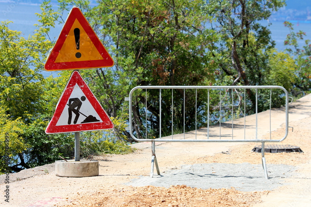 Start of closed road construction site with two heavily used dilapidated warning road works ahead road signs next to protective metal fence blocking access to construction site surrounded with dense t