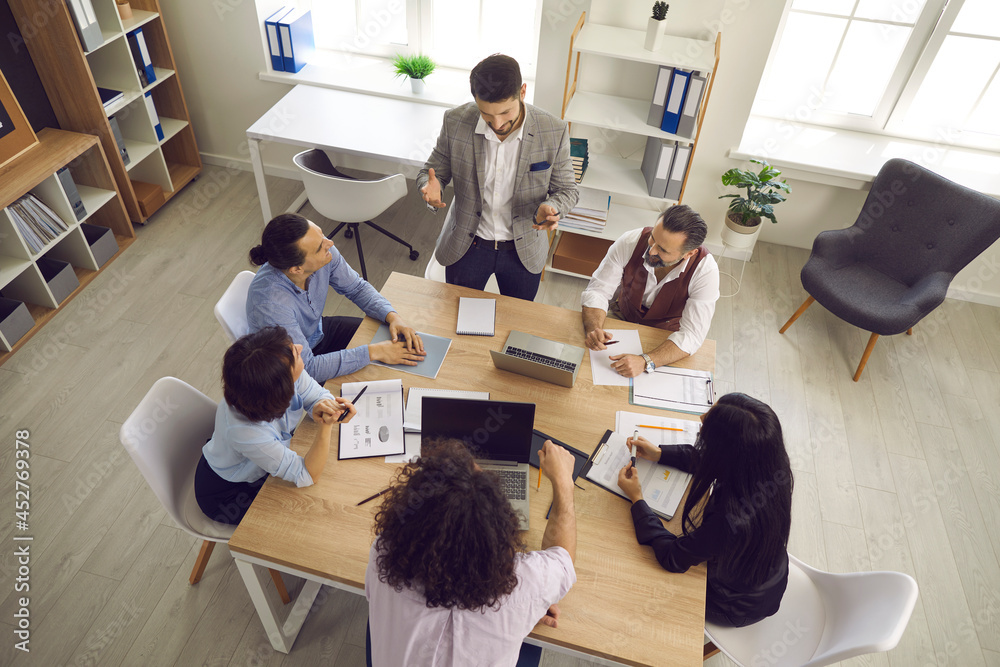 Team of businesspeople having a discussion in a meeting. Group of young and mature business teammates sitting around table in office interior and listening to manager. View from above, high angle shot - obrazy, fototapety, plakaty 