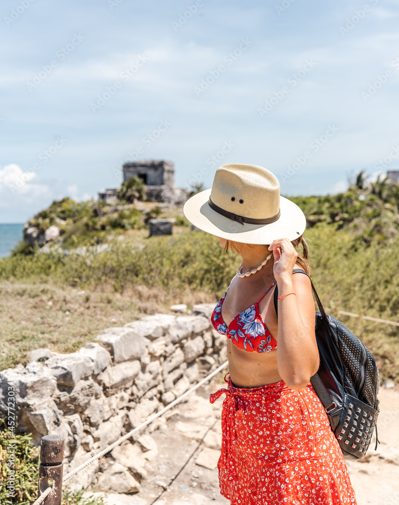 young hispanic woman traveler with hat and backpack looking from above ancient ruins on the coast of the caribbean