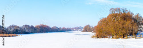 Winter landscape with trees on the banks of the river covered with ice and snow on a sunny day, panorama