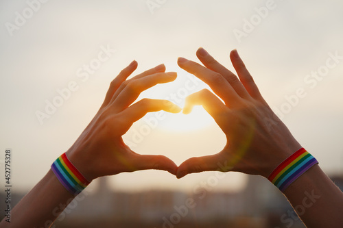 Fototapeta Naklejka Na Ścianę i Meble -  Gay pride, love and marriage concept. Hands with gay pride LGBT rainbow flag wristband making heart sign at sunset sky