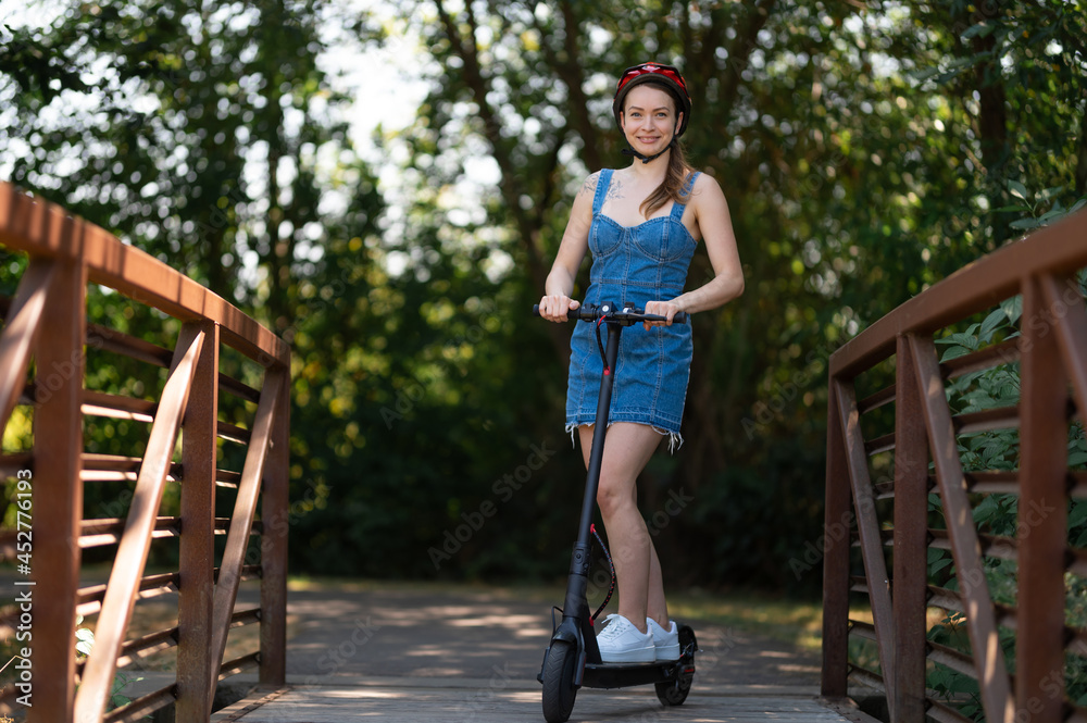 A young blonde girl rides an electric scooter over a wooden bridge. Beautiful nature. Large green trees. Active rest, healthy lifestyle, modern eco-friendly transport.