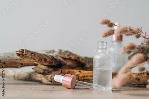 Transparent glass bottle with pipette, scincare serum or essential oil, dried flowers and old wood branch on wooden table. Natural organic cosmetics. photo