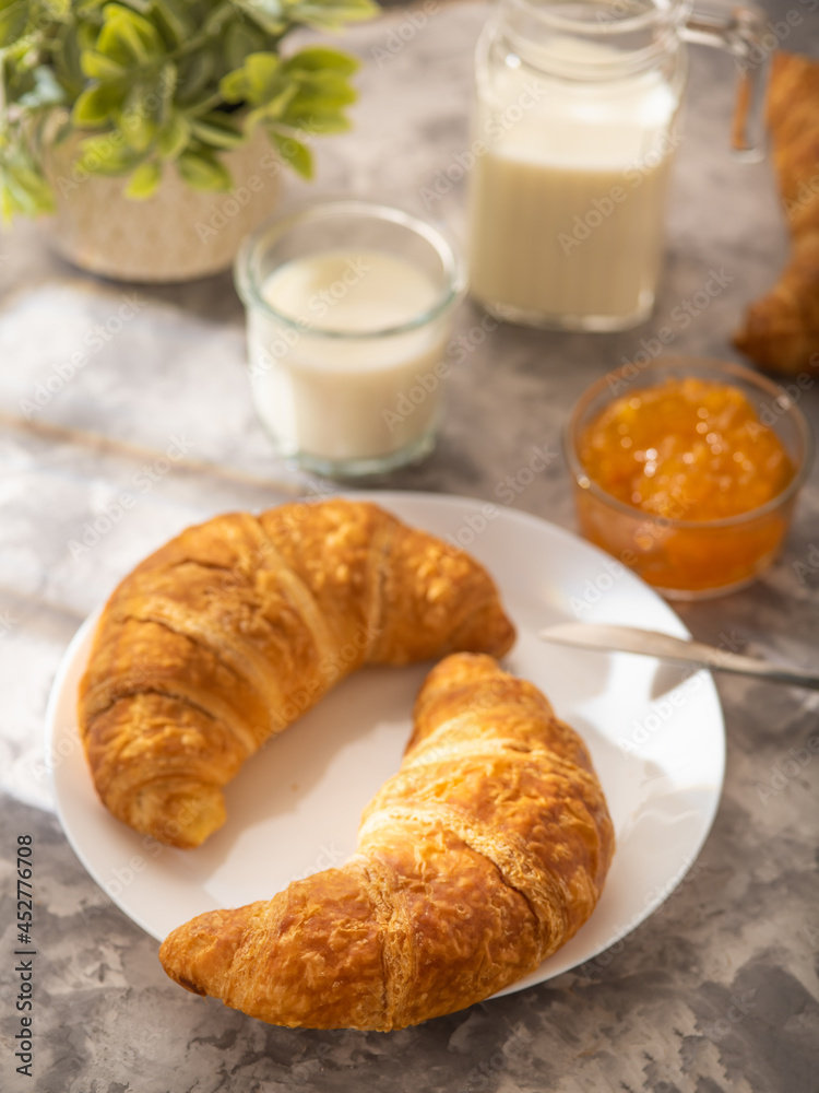 Classic French breakfast - two ruddy croissants on a white faience plate, milk and orange jam. Light background. Low angle view. Restaurant, cafe, bakery, home cuisine, menu design. Classic French breakfast - two ruddy croissants on a white faience plate, milk and orange jam. Light background. Low angle view. Restaurant, cafe, bakery, home cuisine, menu design.