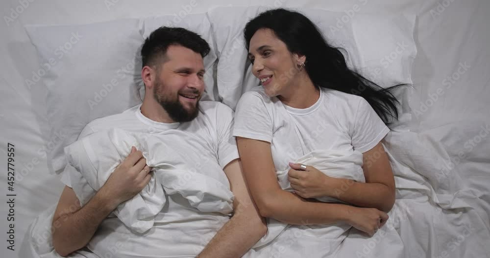 Top view of Caucasian couple laughing and talking in bed with white sheets. High angle shooting. Couple in their 30s talking in bed 4k